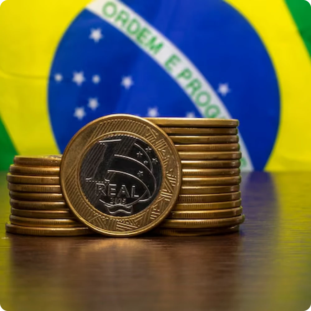 Foto de moedas de um real empilhadas com uma bandeira do brasil ao fundo